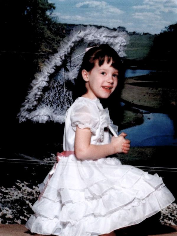A picture of Katie White as a little girl. She is wearing a ruffly white dress and she is holding a lace umbrella. She has brown hair and a sweet smile.